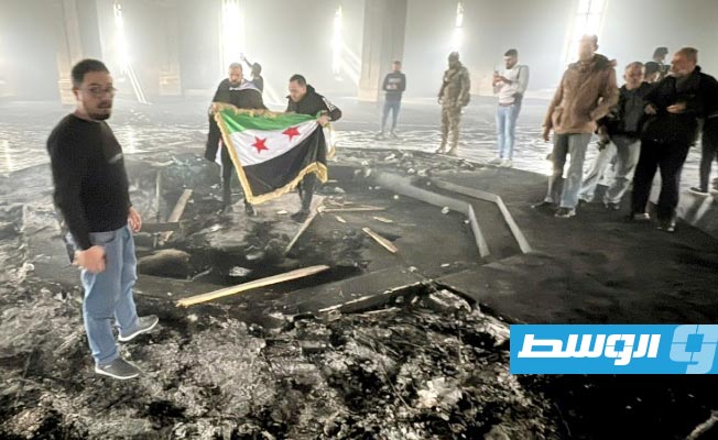 عنصران من المعارضة يحملان علم سورية الجديد فوق قبر الرئيس السوري الراحل حافظ الأسد المحترق في ضريحه بقرية القرداحة، الأربعاء 11 ديسمبر 2025. (أ ف ب)