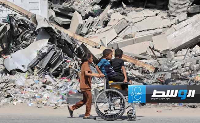 فتاة فلسطينية تدفع أشقائها فوق كرسي متحرك أمام مباني منهار في غزة جراء القصف الإسرائيلي. (الإنترنت)
