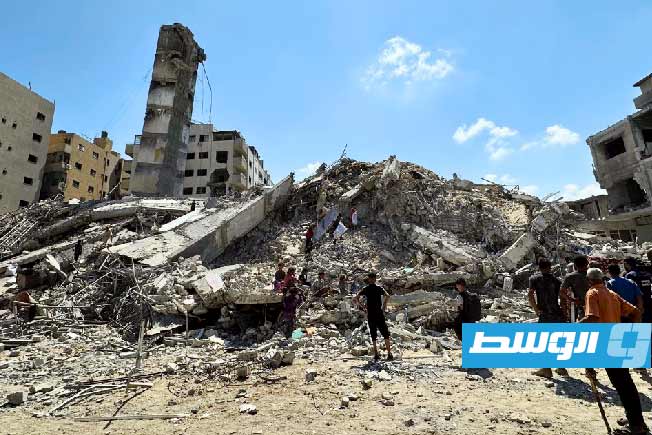 فلسطينيون يقفون بالقرب من أنقاض المباني التي دمرها الاحتلال. (أونروا)
